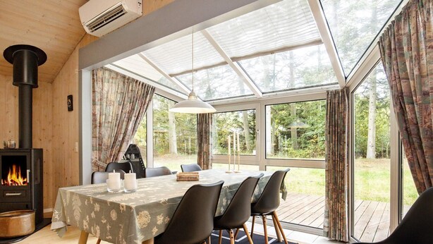 A dining area with a table set for four, a wood-burning stove, and large windows offering a view of the outdoors. - Holiday Home in Saltum (Saltum)