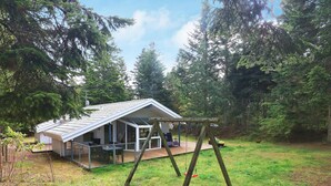A modern house with a glass extension, a wooden deck, and a swing set in a forested area. - Holiday Home in Saltum (Saltum)