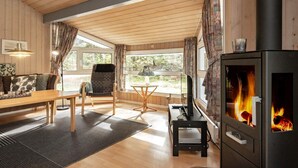 A cozy living room with a wooden floor, a sofa, a dining table, a TV, and a wood-burning stove. - Holiday Home in Saltum (Saltum)