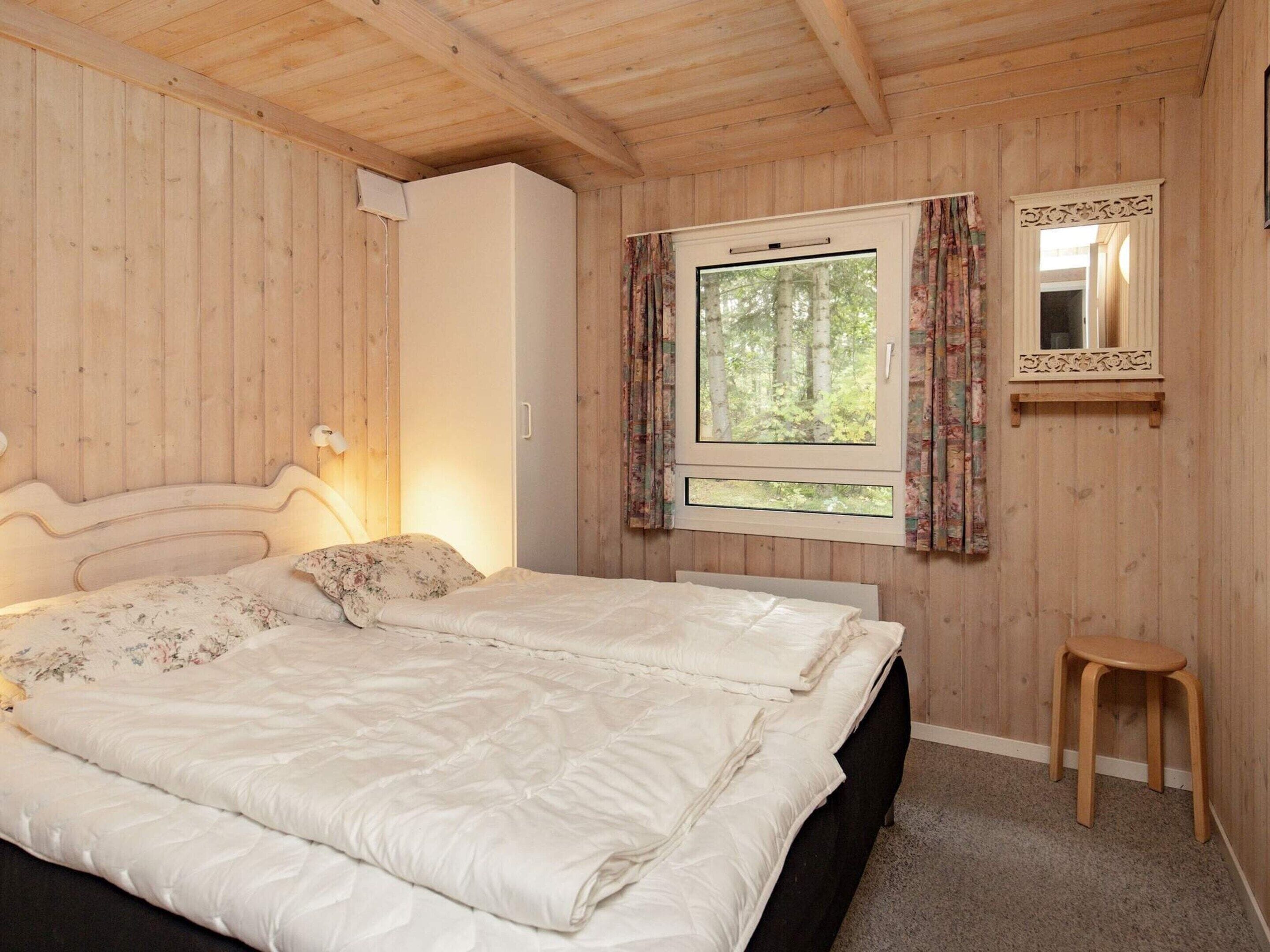 Un dormitorio con cama, ventana con cortinas, espejo y taburete de madera.