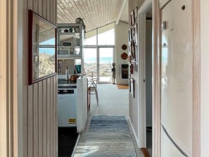 A narrow hallway with a view of the beach, a dining area with a table and chairs, and a kitchen area with cabinets and a sink. - Holiday Home in LÃ¸kken (Lokken)