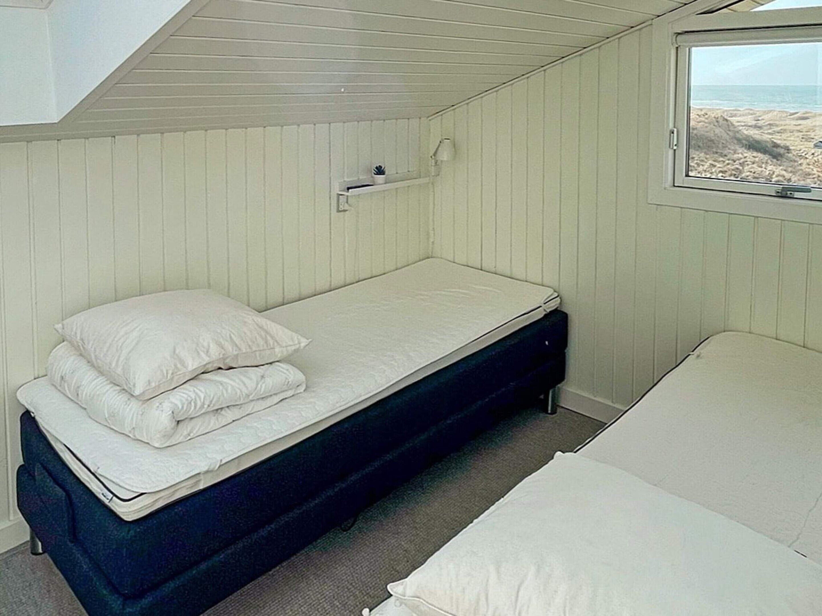 A single bed with a mattress and pillows, a bedside table with a lamp, and a window with a view of the beach.