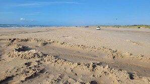 Una playa amplia y abierta con un coche blanco estacionado en la arena, un cielo azul despejado y una vista distante del océano.