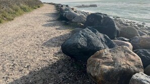 Un sentier de gravier longe une côte rocheuse parsemée de gros blocs, et une colline herbeuse se trouve sur la gauche.