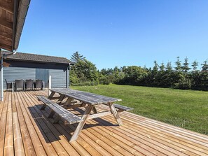 A wooden deck with a picnic table and chairs, a building with a blue exterior, and a grassy yard with trees. - 8 Person Holiday Home in Lokken (Lokken)