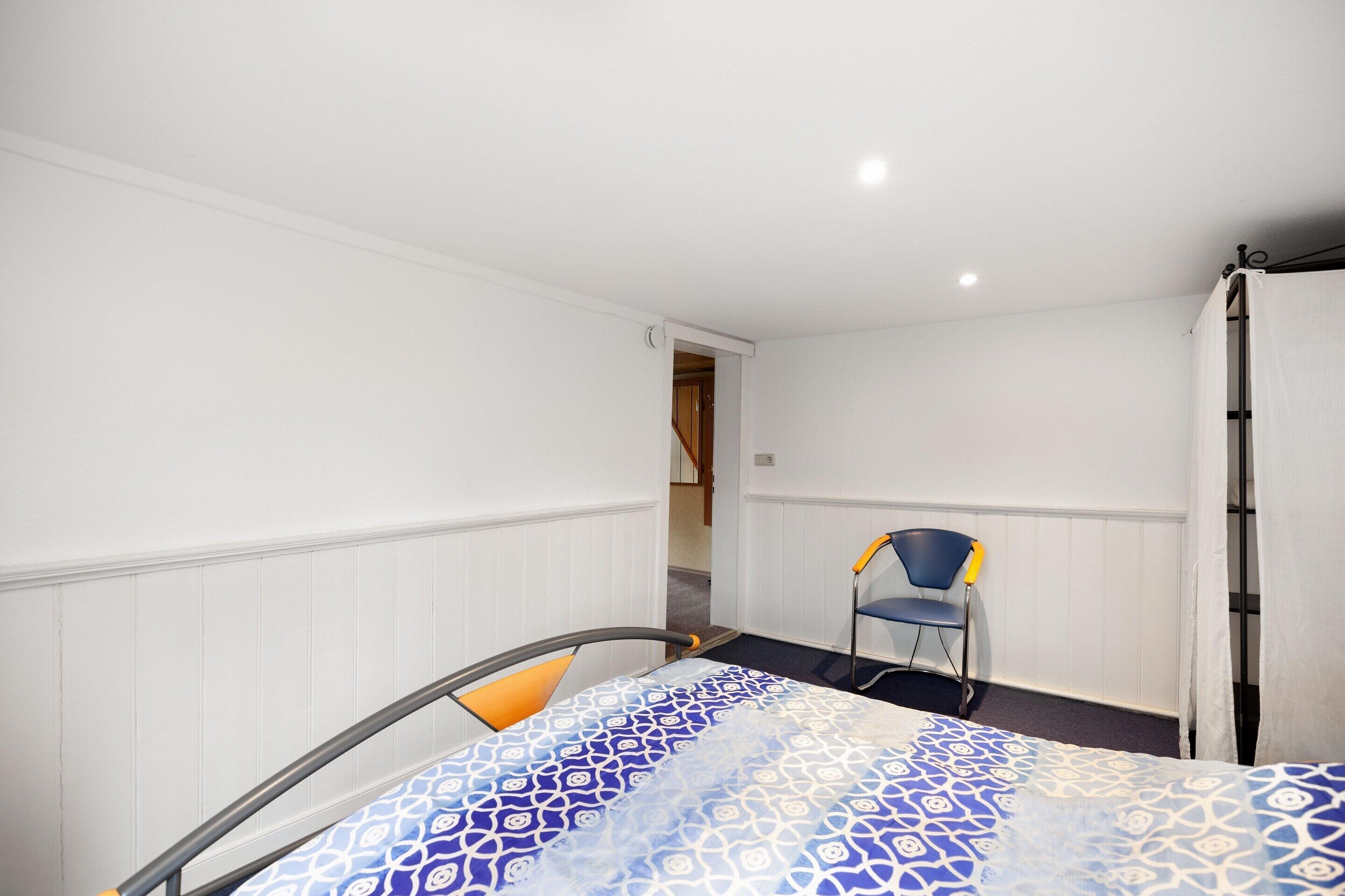 A small, single-bed room with a patterned bedspread, a chair, and a door leading to another room.