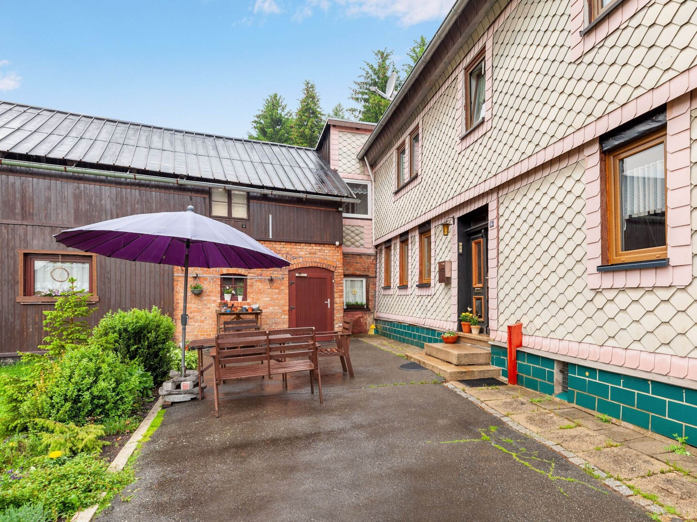 A courtyard with a wooden bench and a purple umbrella, a brick building with a metal roof, and a house with a patterned facade.