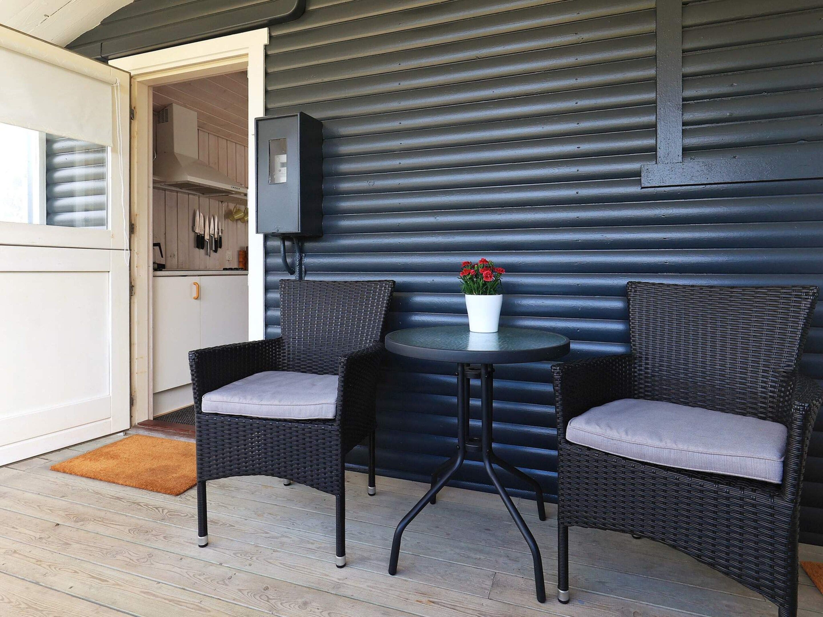 A patio area with two wicker chairs and a small table, a potted plant, and a kitchen visible through an open door.