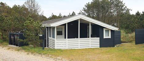A small, single-story cabin with a porch, surrounded by trees and a gravel path.