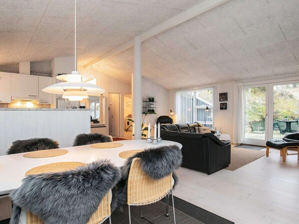 A modern kitchen with white cabinets, a dining area with a white table and wicker chairs, and a black leather sofa in a living area with a large window.