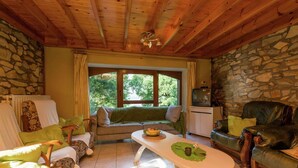 A cozy room with a stone wall, wooden ceiling, a large window, a sofa, a dining table, and a leather couch. - Renovated Farmhouse From 1832 With Beautiful View of Winter Sports Area (Vielsalm)
