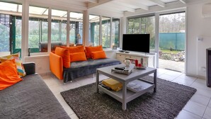 Ein modernes Wohnzimmer mit Sofa, Couchtisch und Fernseher. Große Fenster bieten Blick ins Freie.