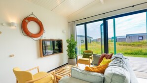 Un salon moderne avec un grand canapé gris, des fauteuils jaunes et une table basse en bois. Un téléviseur encadré en bois est fixé au mur, et on aperçoit une plante en pot.