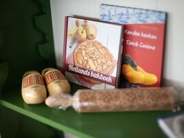 A shelf with two cookbooks, one titled 'Holland's Bakboek' and the other 'Dutch Cuisine'. There are also three small jars with labels and a rolling pin.