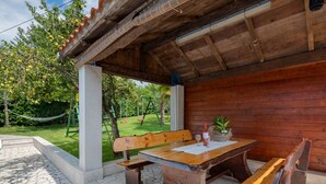 An outdoor wooden table and benches under a covered area with a view of a garden and swings. - Holiday Home in Lindar With Pool (Pazin)