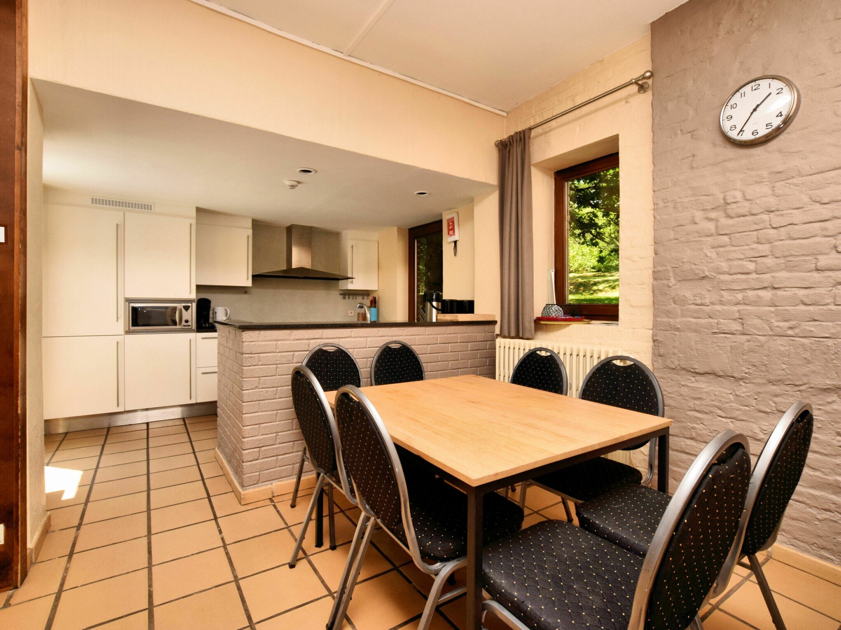 Een compacte keuken met een eethoek, inclusief een tafel en stoelen, een klok aan de muur, en een raam met uitzicht op groen.