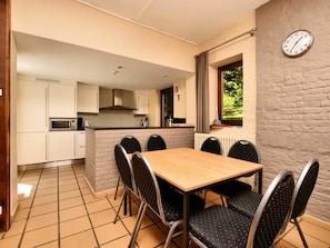 A compact kitchen with a dining area, including a table and chairs, a clock on the wall, and a window with a view of greenery.