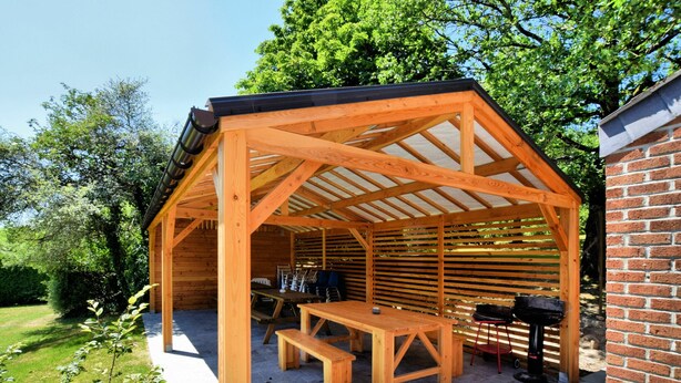 Una pérgola de madera con una mesa y bancos, rodeada por un muro de ladrillo y un jardín.