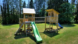 Un parque infantil de madera con toboganes y estructuras para escalar, rodeado de césped y árboles.