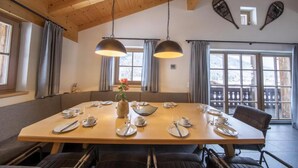 Un'area pranzo con un grande tavolo in legno apparecchiato per quattro persone, due lampade a sospensione e vista all'esterno tramite ampie finestre.