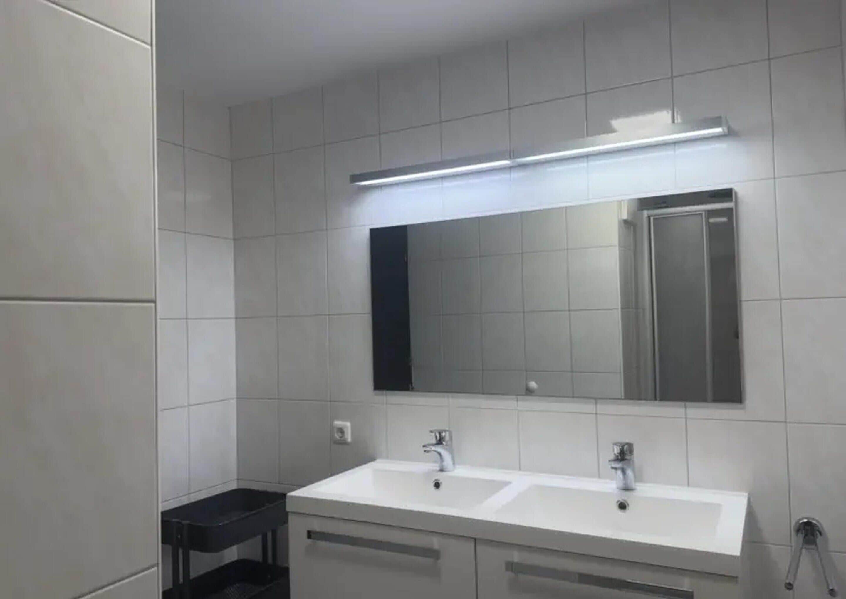 A modern bathroom with white tiled walls, a large mirror, and two sinks.