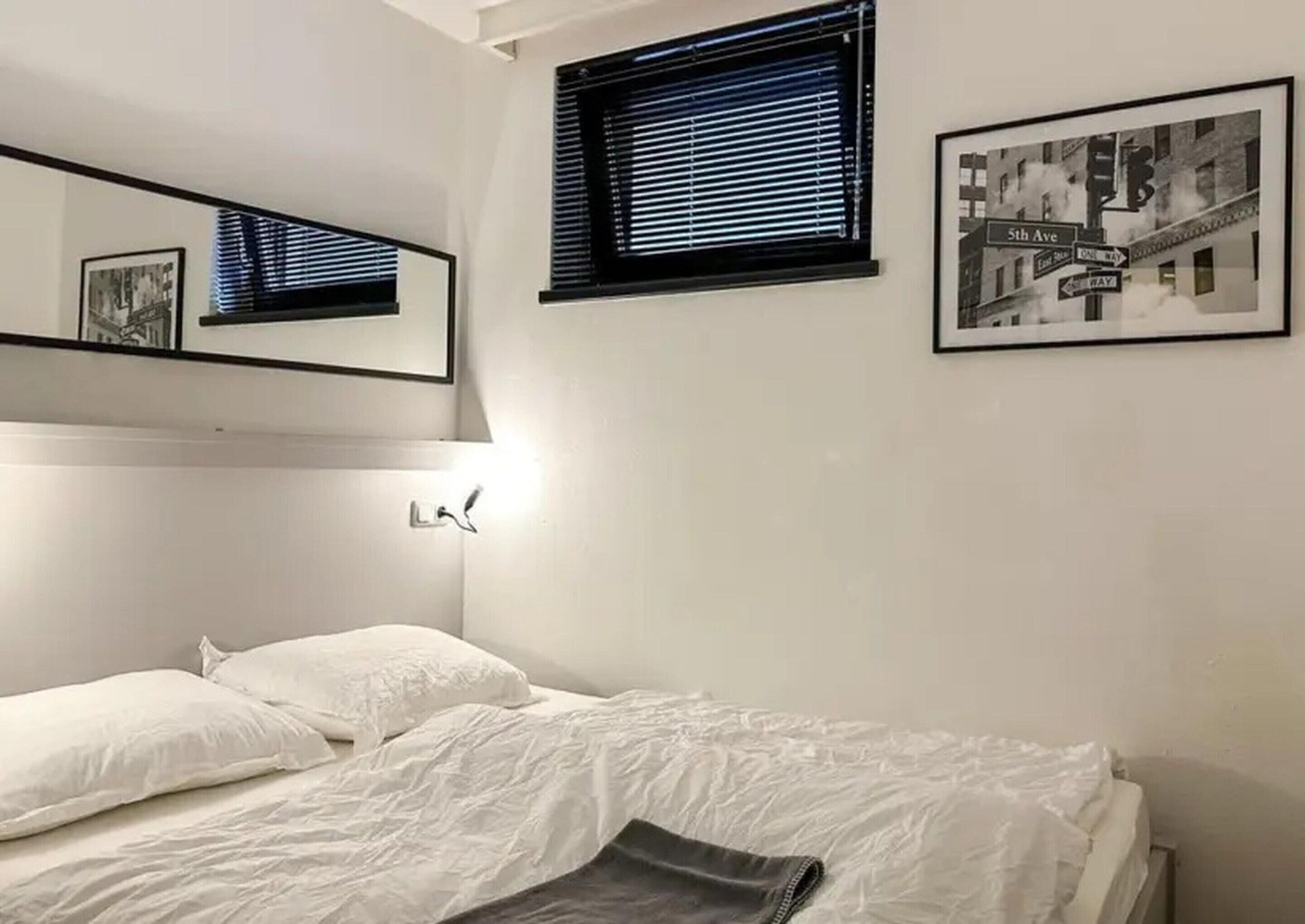 Quarto com uma cama de solteiro, janela com persianas, luminária na parede e um quadro emoldurado.