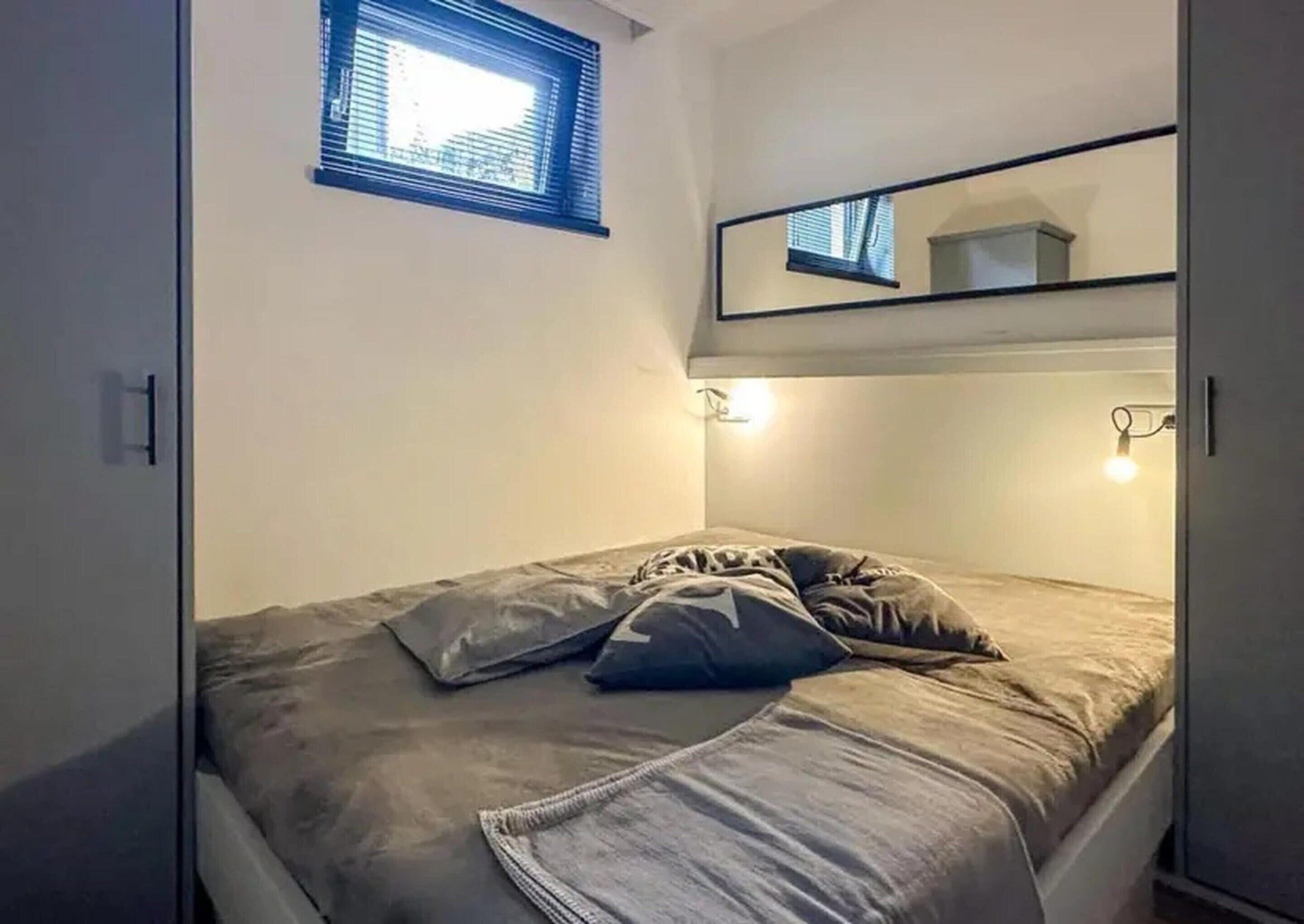 A single bed with a gray mattress and pillows, a small window with blinds, and a mirror above the bed.
