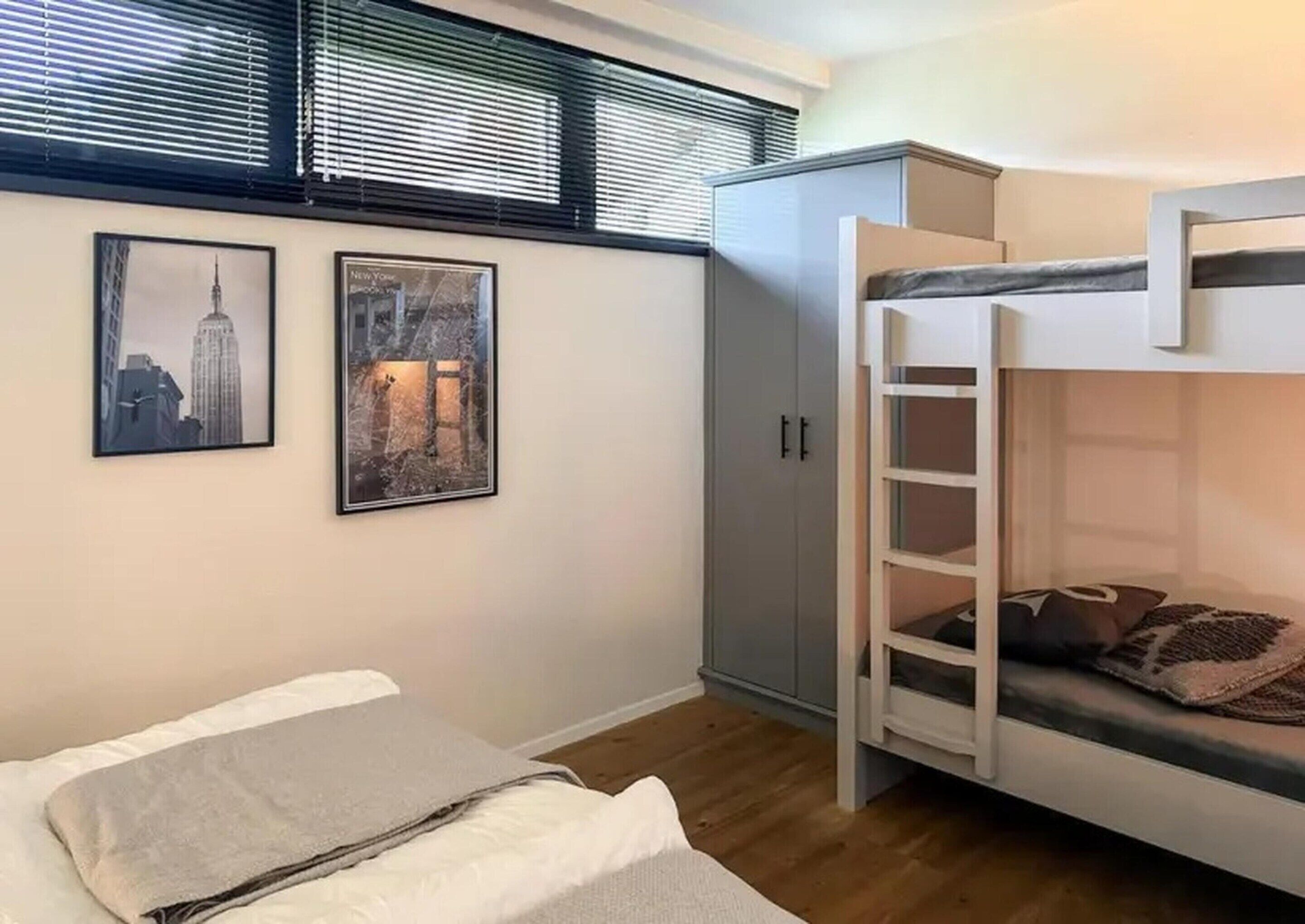 A bunk bed room with a grey wardrobe, two framed pictures on the wall, and a window with blinds.