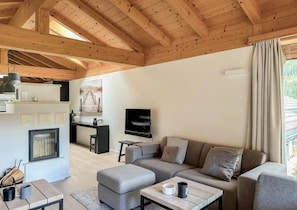 Un salon moderne avec un canapé, un pouf et une table basse. On y trouve une cheminée, un téléviseur fixé au mur, ainsi qu’un coin cuisine équipé d’une cuisinière et de placards.