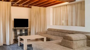 Una sala de estar moderna con techo de madera, televisor de pantalla plana, sofá color beige, mesa de centro de madera y ventanas del suelo al techo con cortinas.