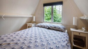 Un dormitorio con techo inclinado, una cama con cubrecama estampado, dos mesitas de noche con lámparas, una ventana con vistas a la vegetación y un reloj sobre una de las mesitas.