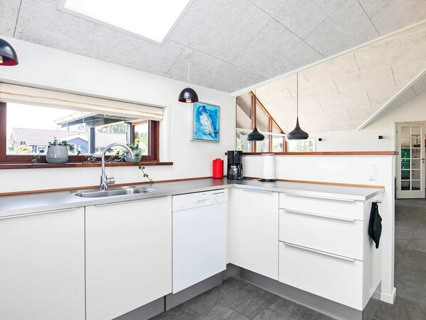 A modern kitchen with white cabinets, a stainless steel sink, and a window overlooking a residential area. - Holiday Home in Løkken (Lokken)