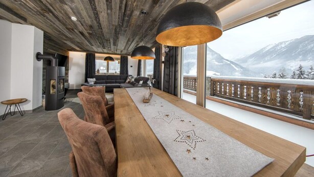 Un espace de repas moderne avec une longue table en bois, des chaises à revêtement marron et des luminaires suspendus. Une grande fenêtre offre une vue sur des montagnes enneigées.
