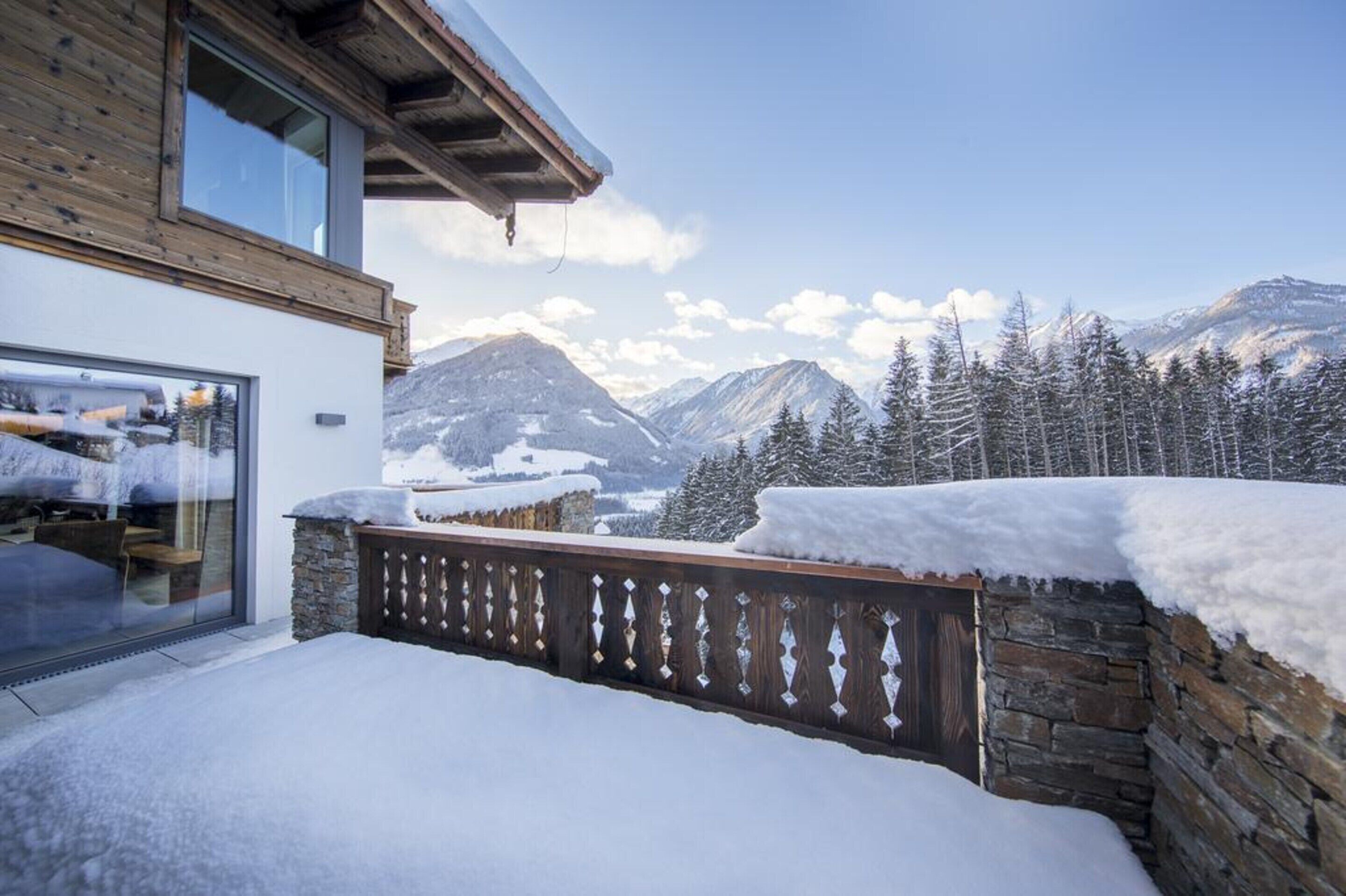 A modern building with a stone wall and wooden deck, overlooking a snowy mountain landscape.