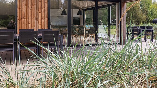 A modern outdoor seating area with wooden tables and chairs, surrounded by tall grasses and a wooden structure with glass doors. - Holiday Home in Sæby (Sæby)