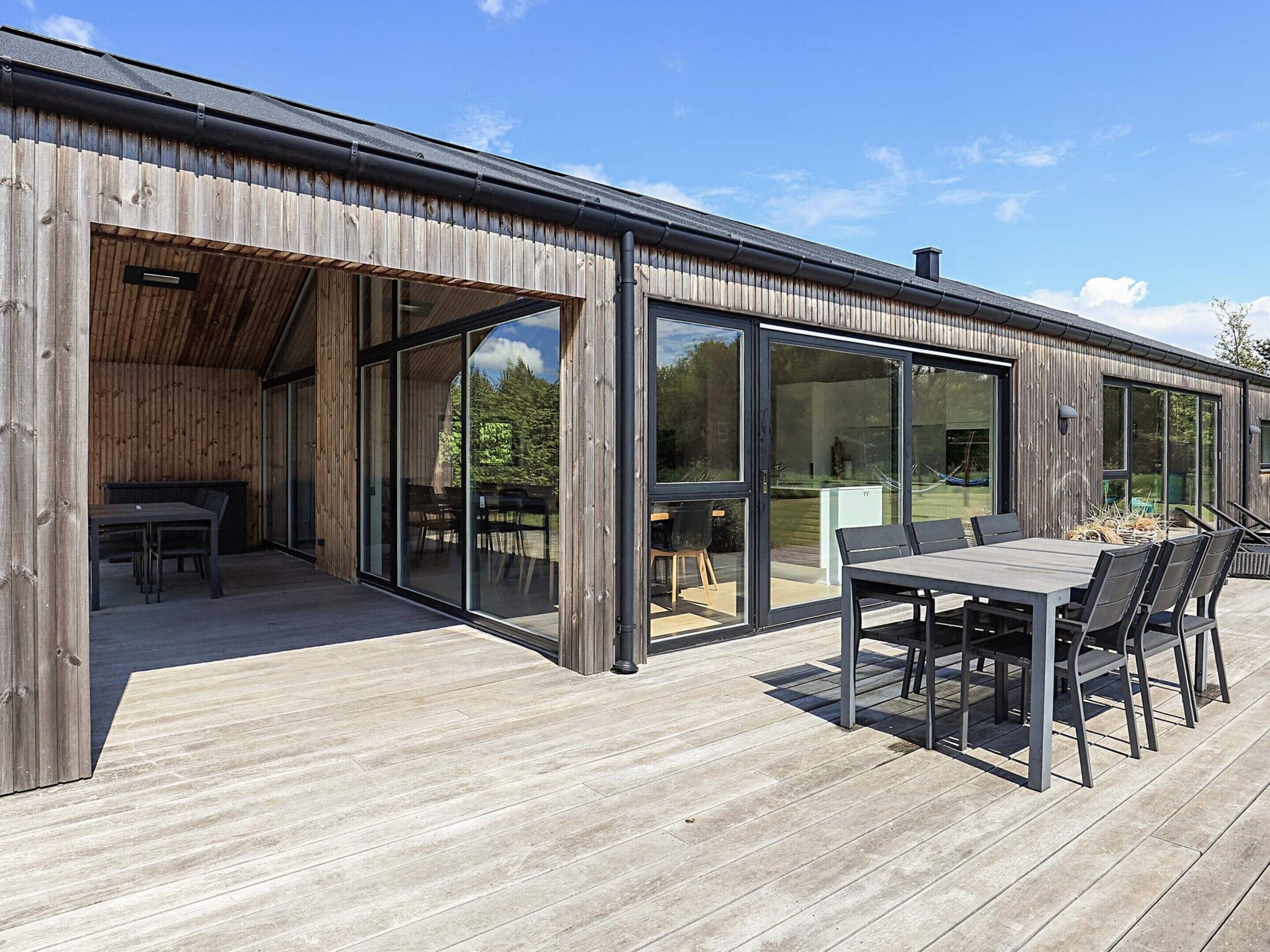 Ein modernes Gebäude mit großen Glasfenstern und -türen, einer Holzterrasse und Sitzgelegenheiten im Freien.