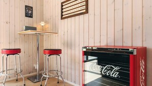 A room with wooden panel walls, a red Coca-Cola vending machine, two bar stools, a table with a lamp, and a poster on the wall. - Holiday Home in Sæby (Sæby)