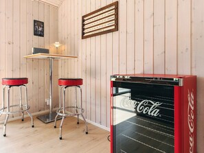 A room with wooden panel walls, a red Coca-Cola vending machine, two bar stools, a table with a lamp, and a poster on the wall. - Holiday Home in Sæby (Sæby)