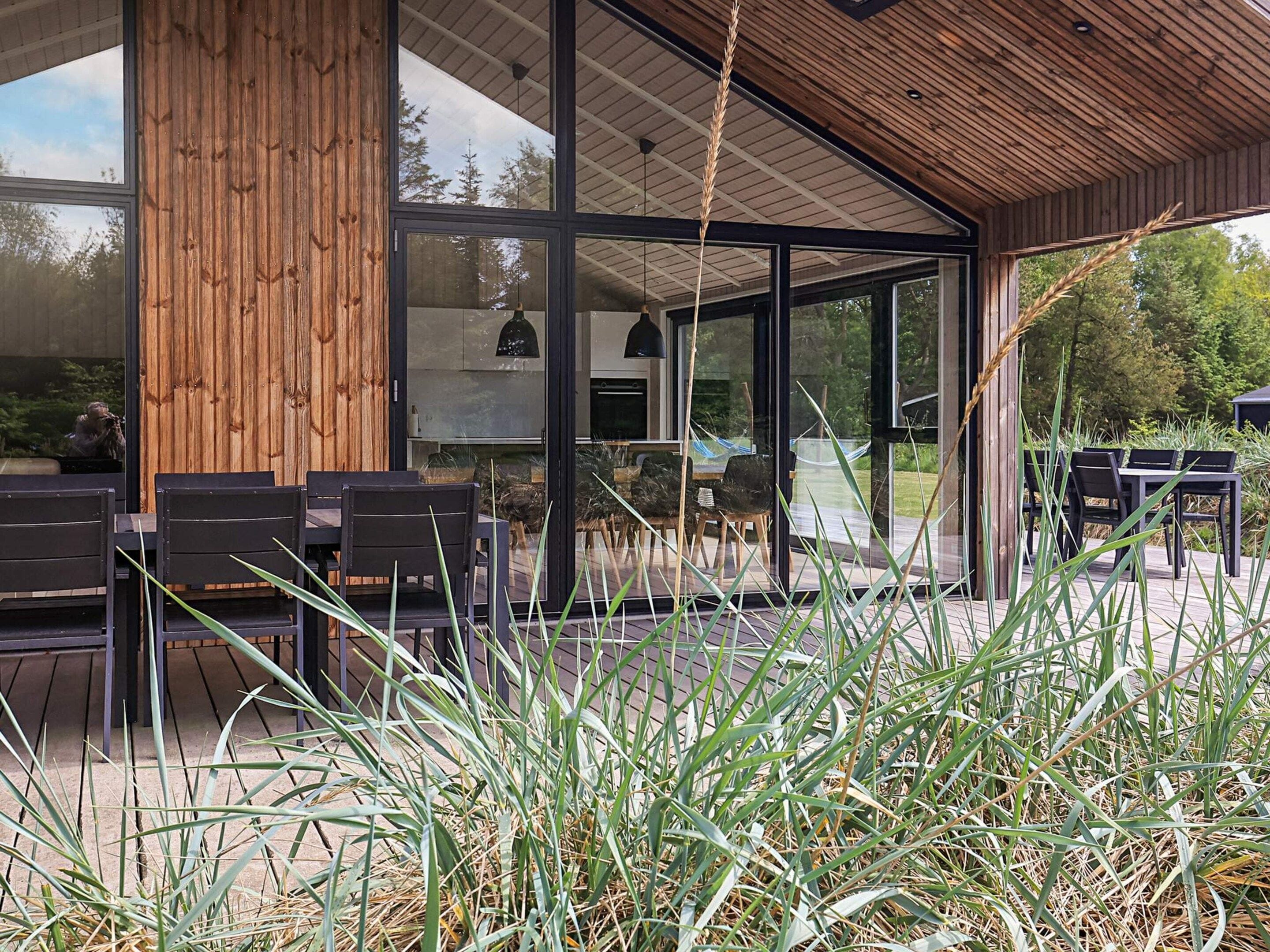 A modern outdoor seating area with wooden tables and chairs, surrounded by tall grasses and a wooden structure with glass doors.