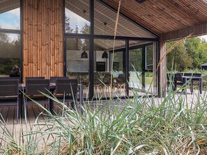 A modern outdoor seating area with wooden tables and chairs, surrounded by tall grasses and a wooden structure with glass doors. - Holiday Home in Sæby (Sæby)