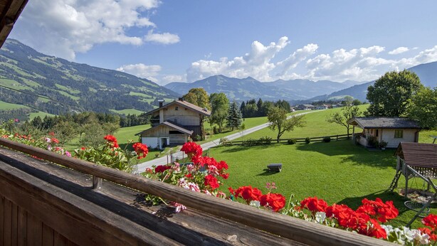 Une vue pittoresque depuis un balcon doté d’une balustrade en bois, donnant sur une vallée verdoyante, avec des maisons et des montagnes en arrière-plan.