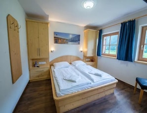 A bedroom with a wooden bed, wooden wardrobe, bedside tables, a window with curtains, and a painting on the wall.