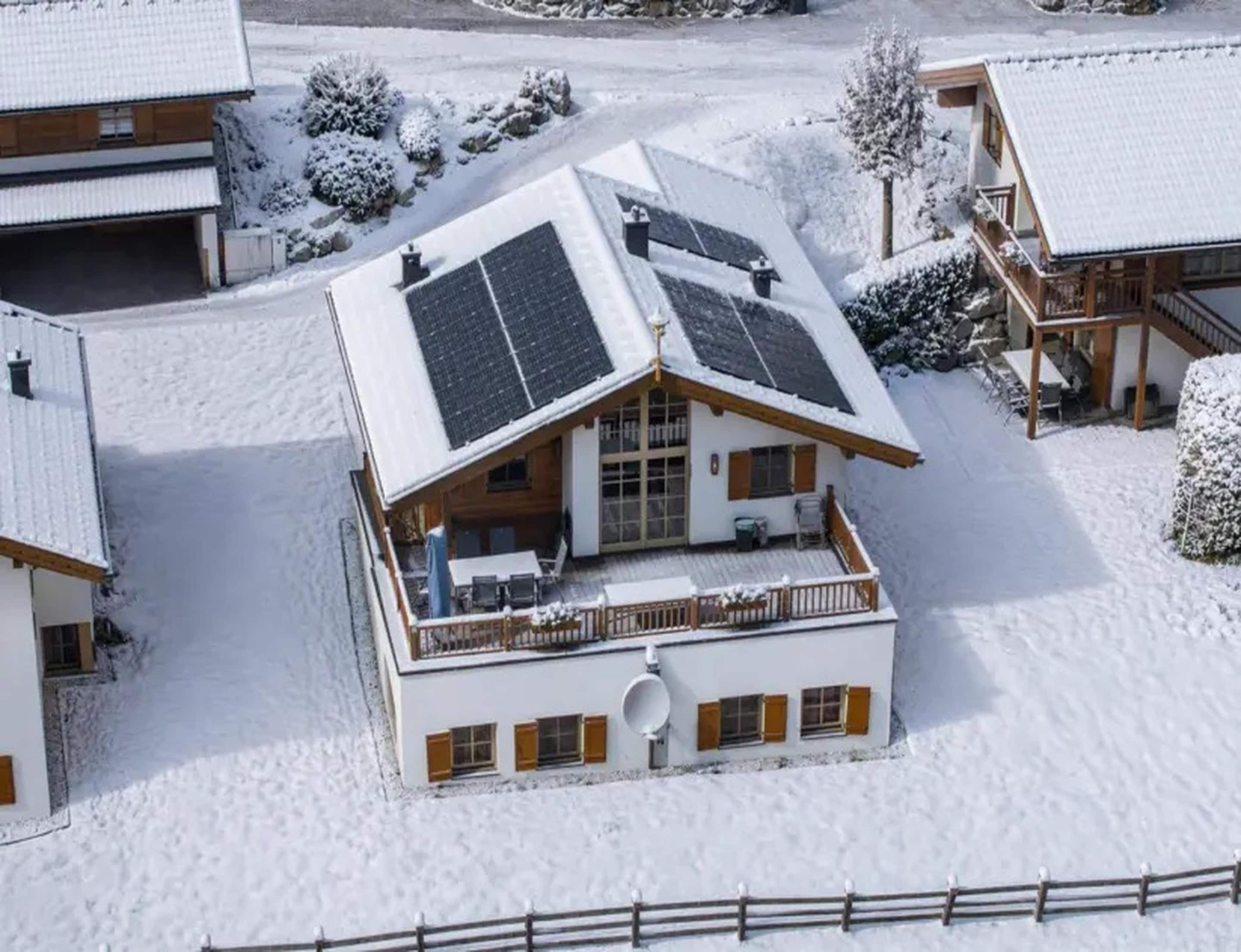 A snow-covered building with a sloped roof, solar panels, and a balcony with outdoor seating.