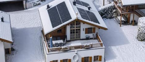 A snow-covered building with a sloped roof, solar panels, and a balcony with outdoor seating.