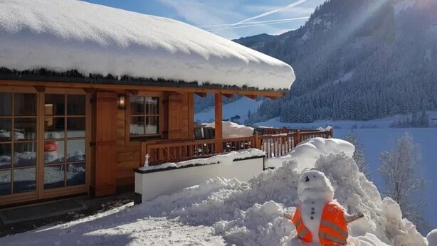 A wooden cabin with a snow-covered roof and a snowman with an orange vest in front. - Cozy Chalet with 4 Bathrooms near Königsleiten (Wald im Pinzgau)