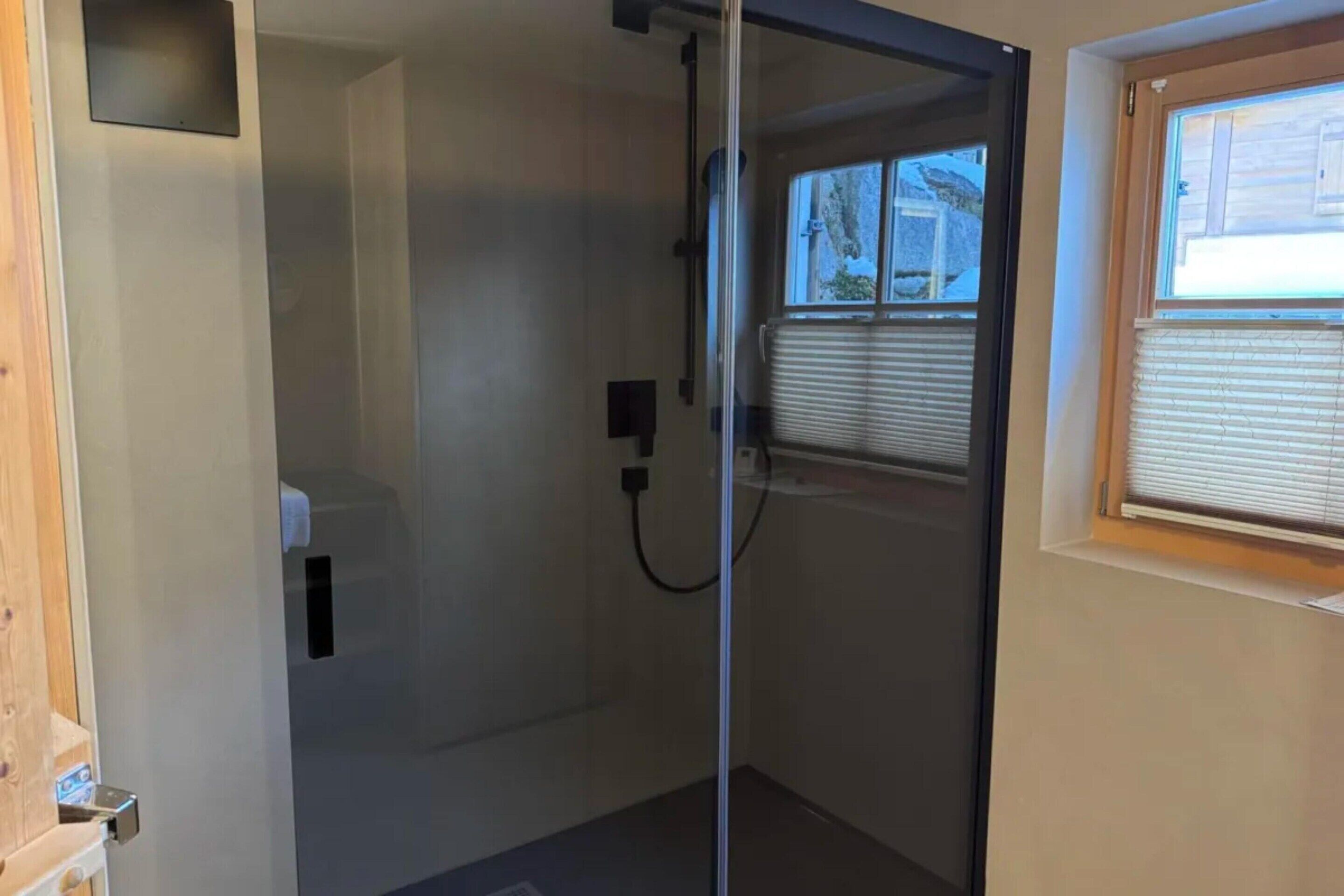 Een douche met glazen wanden, een zichtbare douche slang en een raam met zonwering.