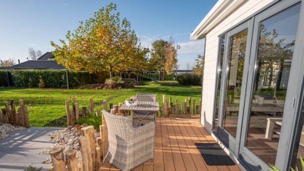 A patio with a wicker table and chair, a wooden fence, and a grassy yard with trees. - Chalet in Goedereede Near North Sea Beach (Goedereede)