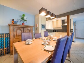 Een eethoek met een houten tafel gedekt voor vier personen, omringd door paarse stoelen. Achtergrond toont een keuken met moderne apparatuur.