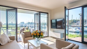 A modern living room with a sofa, chairs, a coffee table, a TV, and a large window with a marina view. - Apartment in Kamperland With Marina Views (Kamperland)