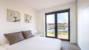 A modern bedroom with a large bed, a nightstand, a lamp, and two wall-mounted paintings. A sliding glass door leads to a balcony with a marina view. - Apartment in Kamperland With Marina Views (Kamperland)